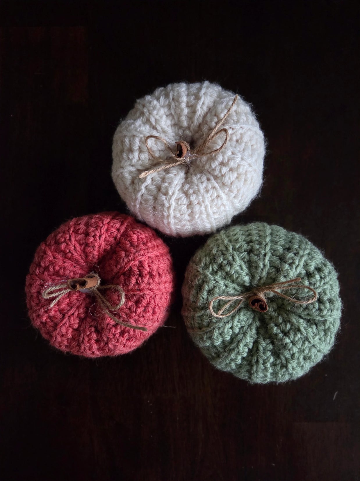 Three knitted pumpkins in cream, red, and green with twine bows on dark background