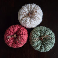 Three knitted pumpkins in cream, red, and green with twine bows on dark background