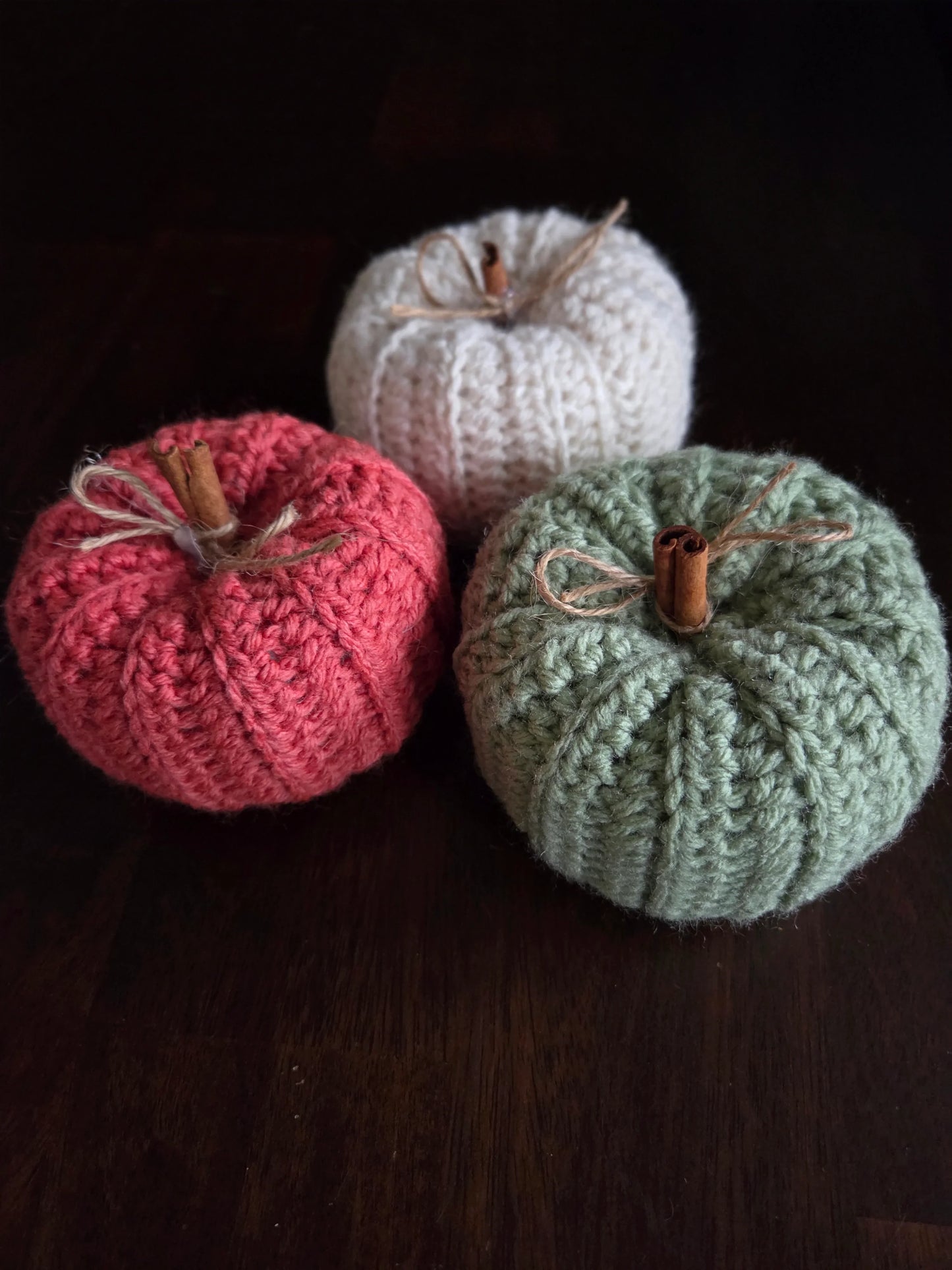Three knitted pumpkins in white, pink, and green with cinnamon stick stems on dark wood