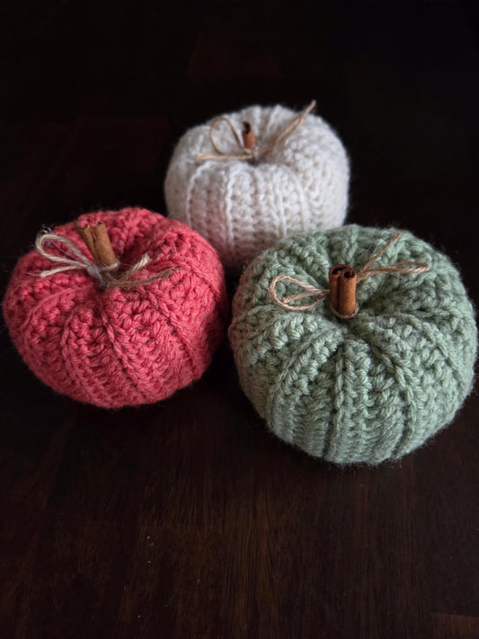 Three knitted pumpkins in white, pink, and green with cinnamon stick stems on dark wood