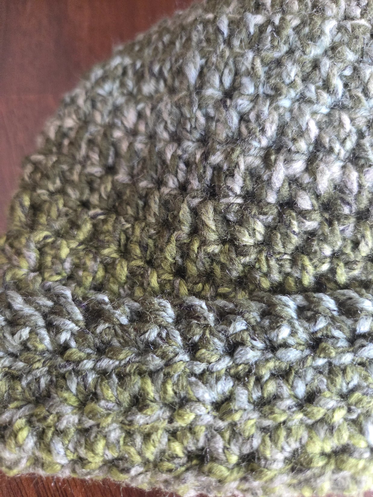 Close-up of a green and gray crochet beanie hat with textured yarn on a wooden surface.