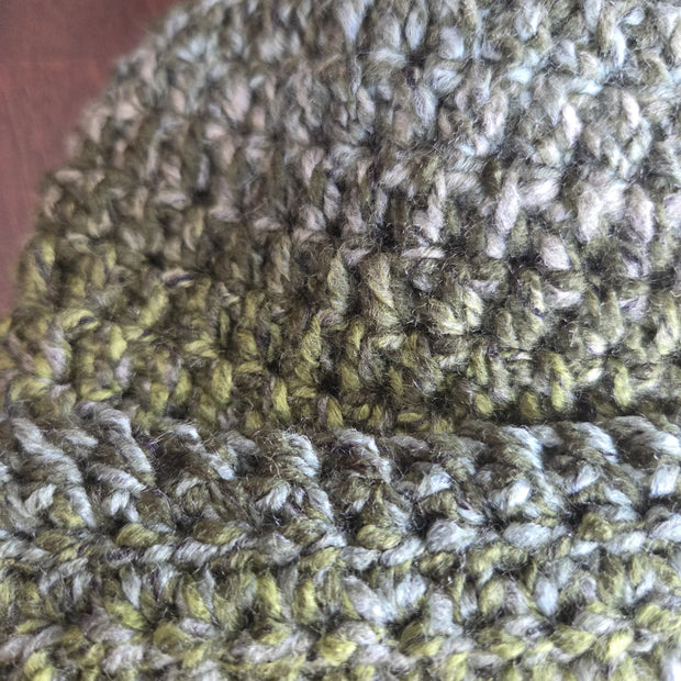 Close-up of a green and gray crochet beanie hat with textured yarn on a wooden surface.