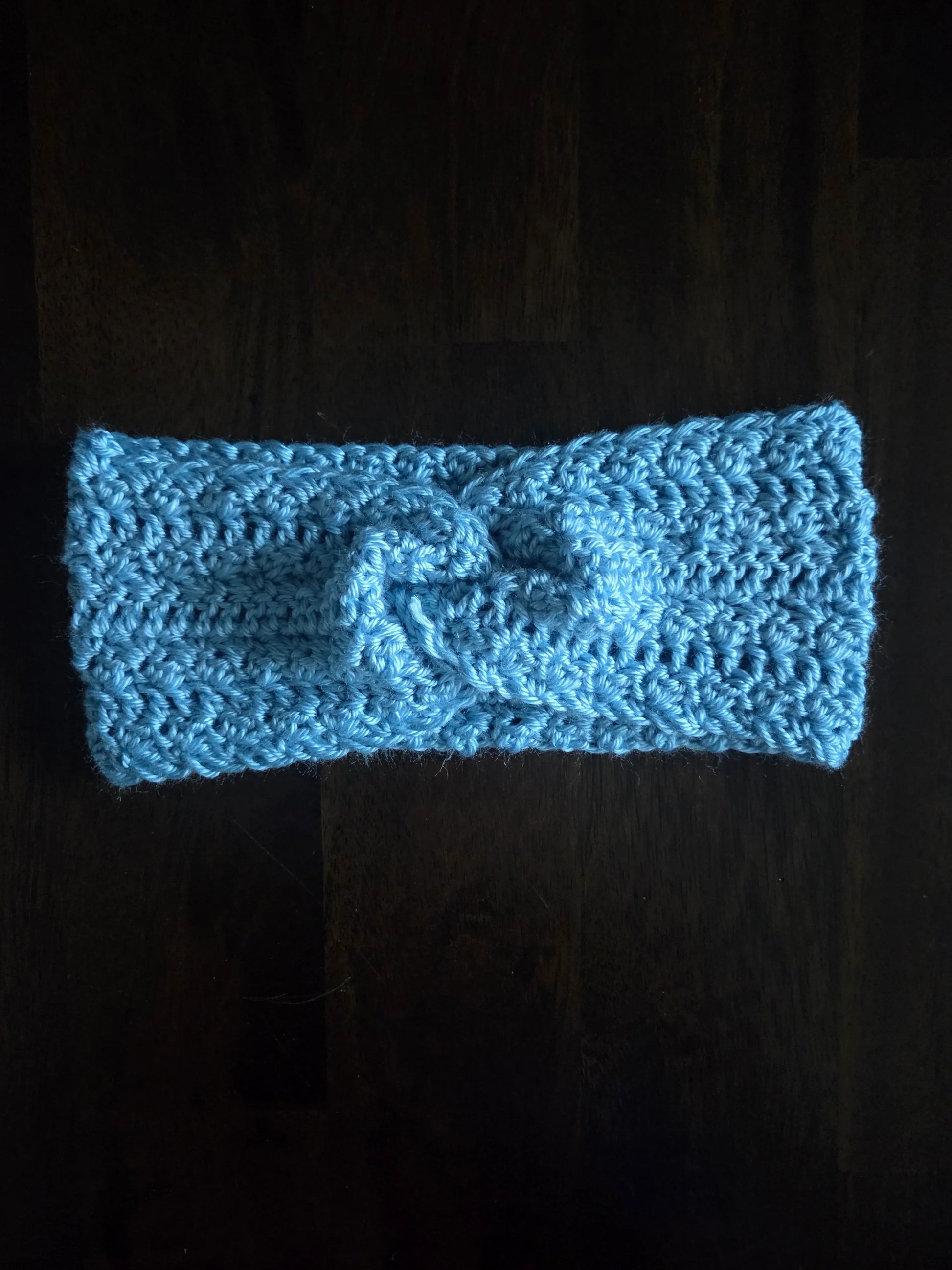 Handmade light blue crochet headband with knot, displayed on a dark wooden surface