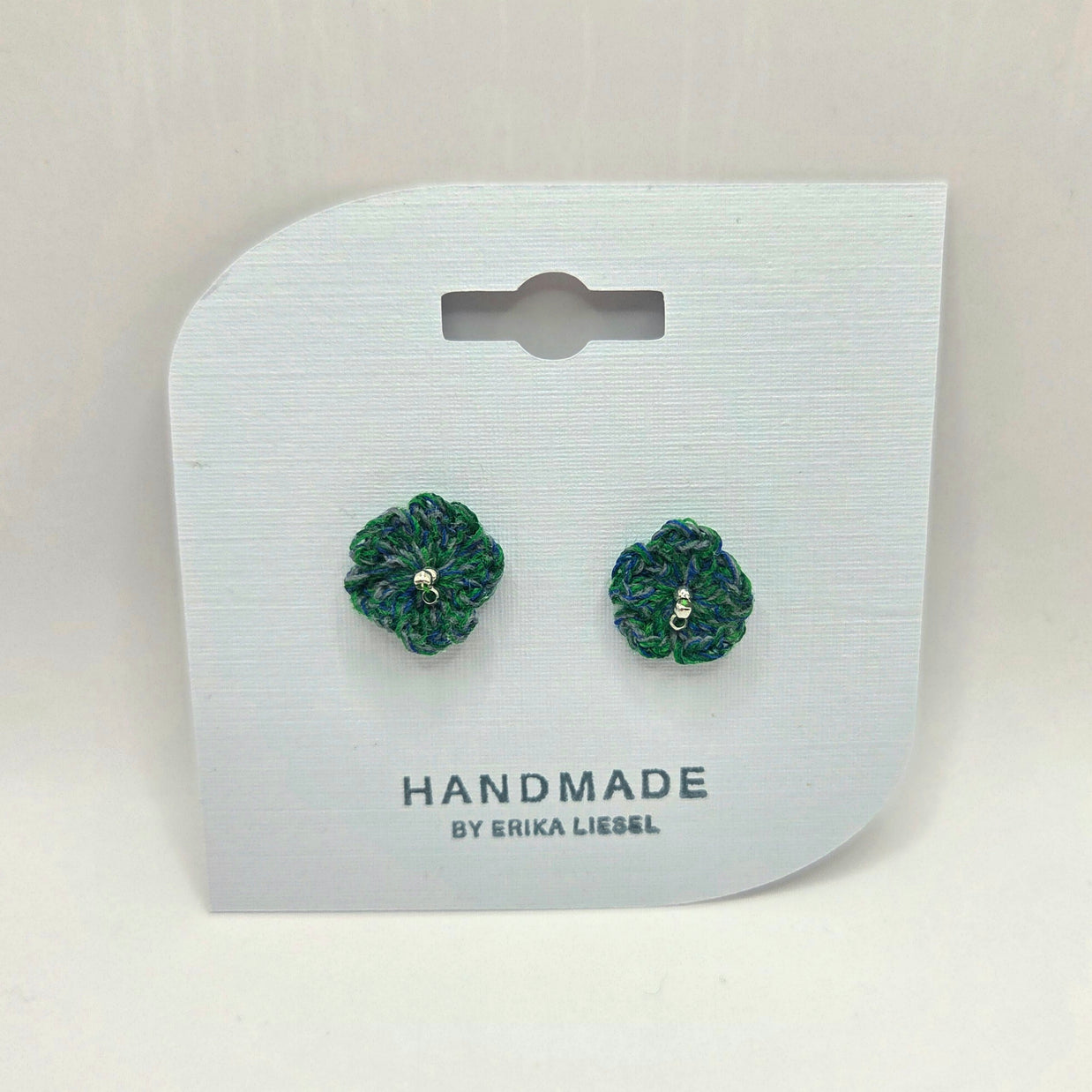 Handmade crochet flower earrings in green and blue, displayed on white card with 'Handmade by Erika Liesel' text.