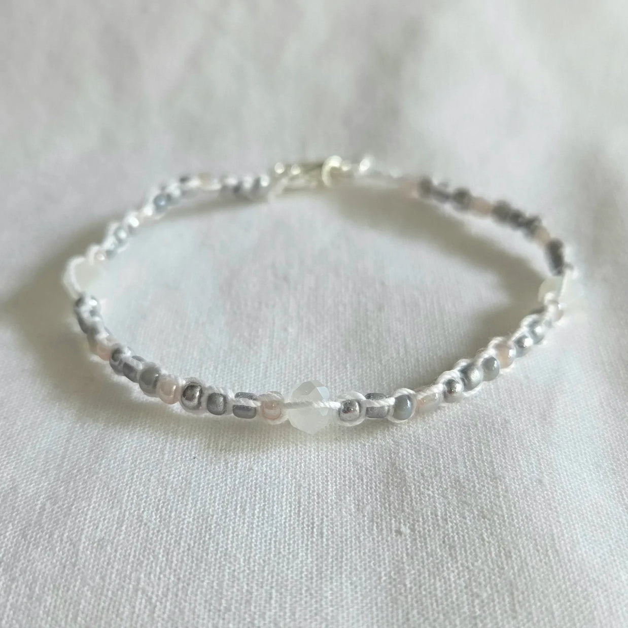 Delicate beaded bracelet with silver, white, and gray beads on white fabric, handmade jewelry gift