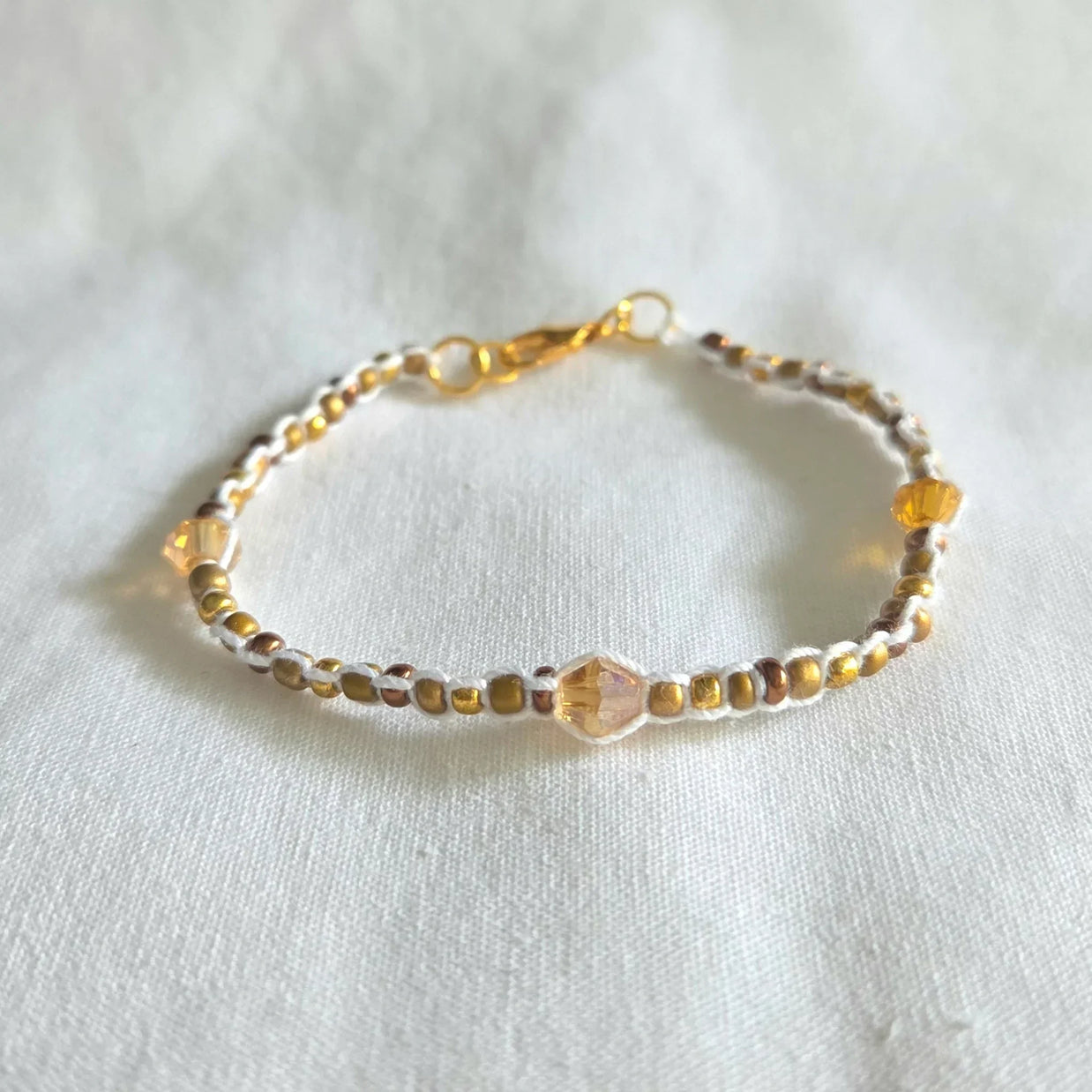 Handmade beaded bracelet with gold, amber, and white beads on white fabric background