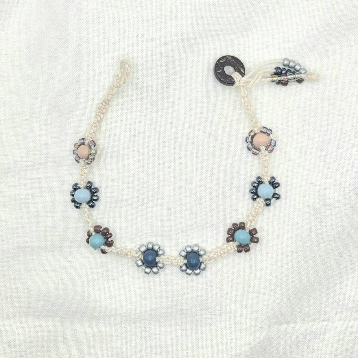Handmade macrame necklace with beaded flower accents in blue, gray, and brown tones on cream cord