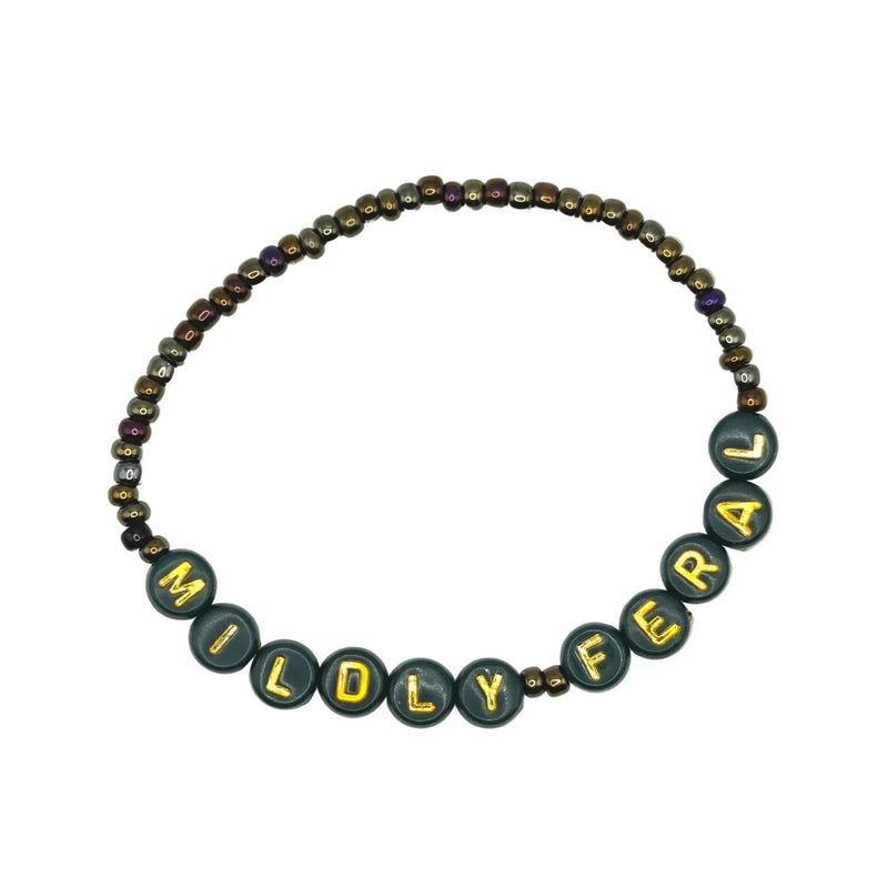 A beaded necklace spelling out 'MILDY FERAL' in dark green letter beads with gold lettering, accented by small metallic bronze and purple beads, on a light gray background.