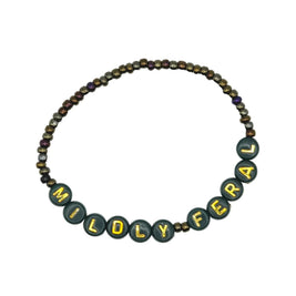 A beaded necklace spelling out 'MILDY FERAL' in dark green letter beads with gold lettering, accented by small metallic bronze and purple beads, on a light gray background.