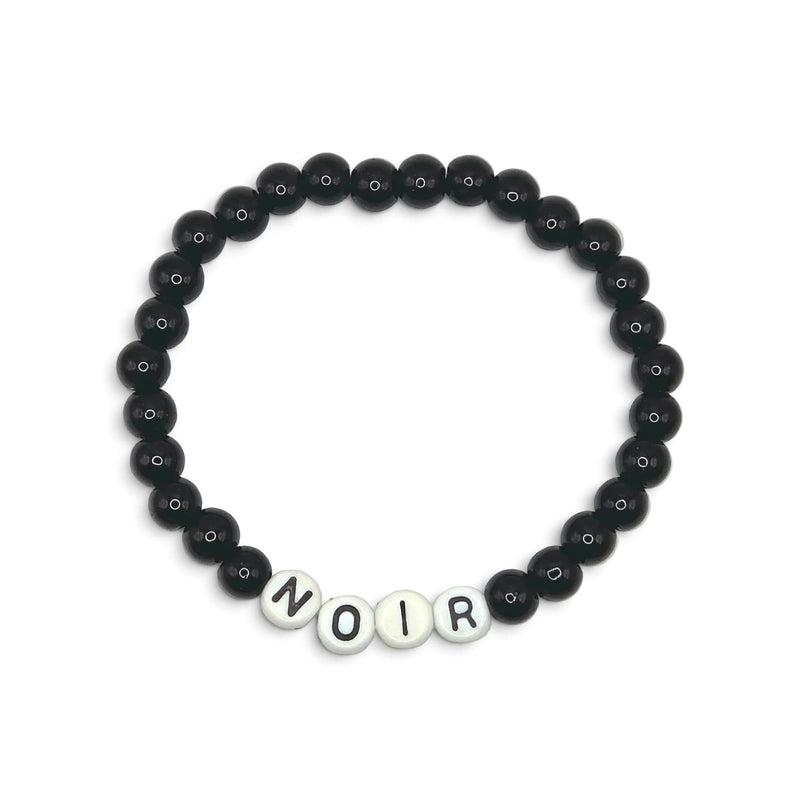 A beaded stretch bracelet featuring matte black round beads and four white letter beads spelling 'NOIR' at the bottom, displayed on a clean white background.