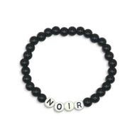 A beaded stretch bracelet featuring matte black round beads and four white letter beads spelling 'NOIR' at the bottom, displayed on a clean white background.