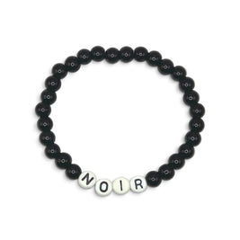 A beaded stretch bracelet featuring matte black round beads and four white letter beads spelling 'NOIR' at the bottom, displayed on a clean white background.