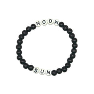 A beaded stretch bracelet on a light gray background, featuring black round beads and white square letter beads spelling 'NOOW SUN'.