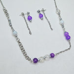 A silver-tone jewelry set featuring a beaded necklace and matching drop earrings, decorated with purple beads, white beads, and clear faceted beads, displayed on a light textured background.