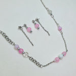 Silver-tone chain jewelry set with pink and white beads, including a necklace, matching drop earrings, and a bracelet, displayed on a light textured fabric background.