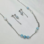 A silver-toned jewelry set featuring a delicate chain necklace, matching drop earrings, and a bracelet, decorated with light blue marbled beads and white pearl-like beads on a textured white fabric background.