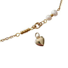 A gold chain bracelet featuring a small dangling heart charm, gold beads, and two white pearl-like beads on a plain light background.