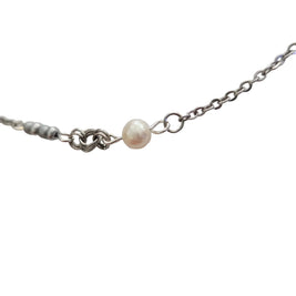 A close-up of a delicate silver chain necklace featuring a single small, lustrous white pearl bead and a short section of textured silver beads on the left, set against a plain light gray background.