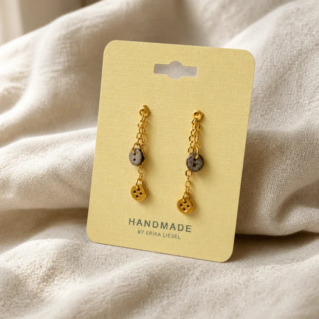 Handmade gold chain earrings with button charms, displayed on a pale yellow card against soft fabric.
