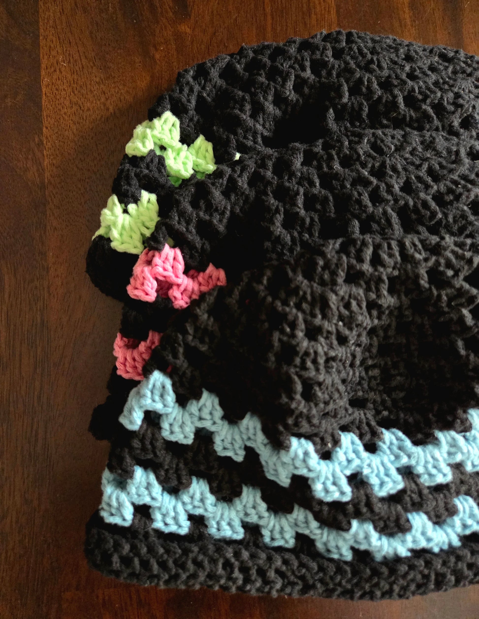 Stack of handmade black crochet hats with green, pink, and blue accent stripes on wood table