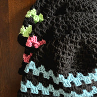 Stack of handmade black crochet hats with green, pink, and blue accent stripes on wood table