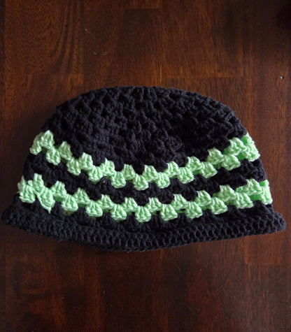 Handmade crochet beanie with black and green stripes on a wood surface.