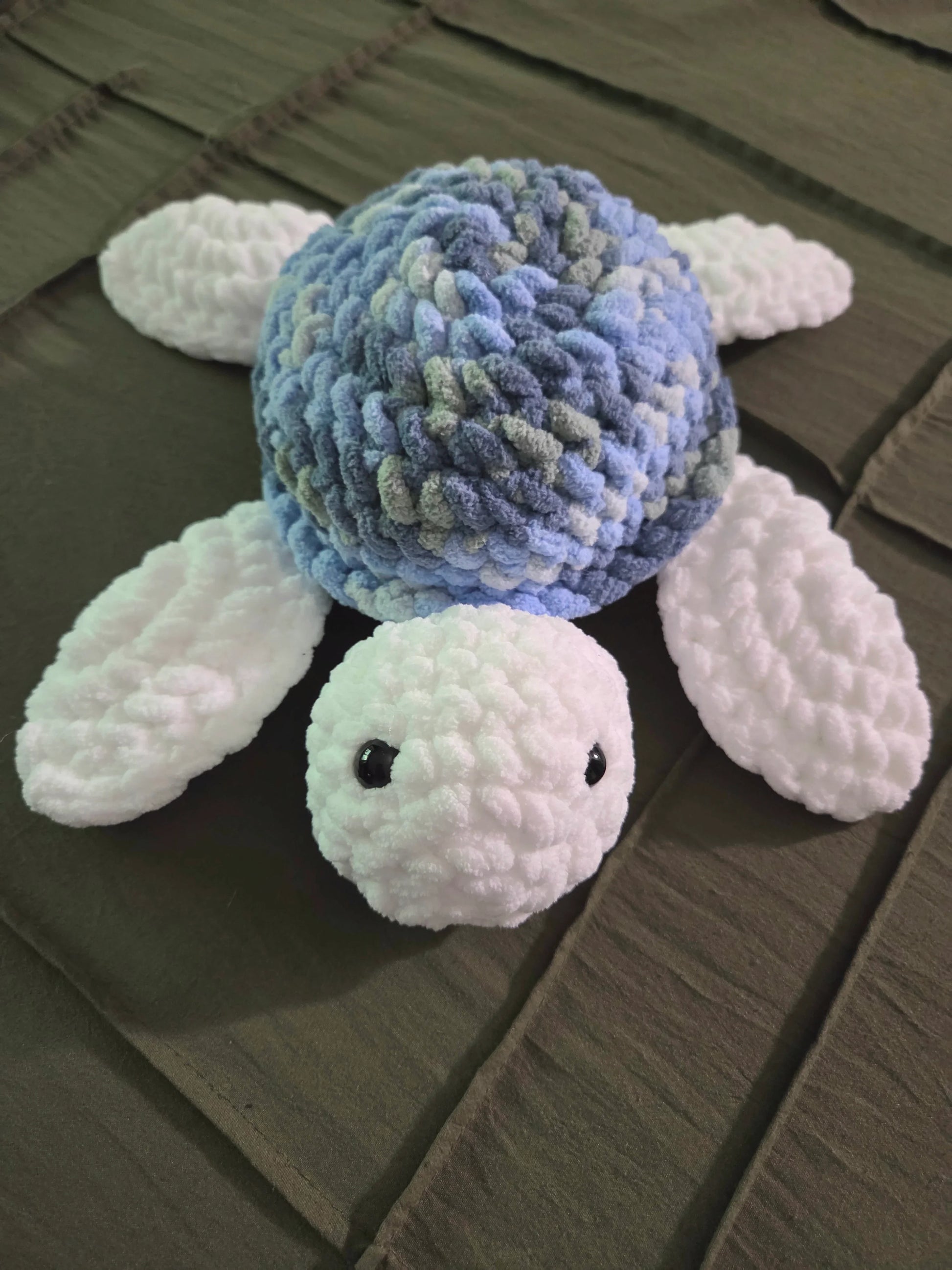 Plush crochet turtle toy with blue shell and white flippers on a dark quilted blanket