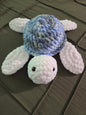 Plush crochet turtle toy with blue shell and white flippers on a dark quilted blanket