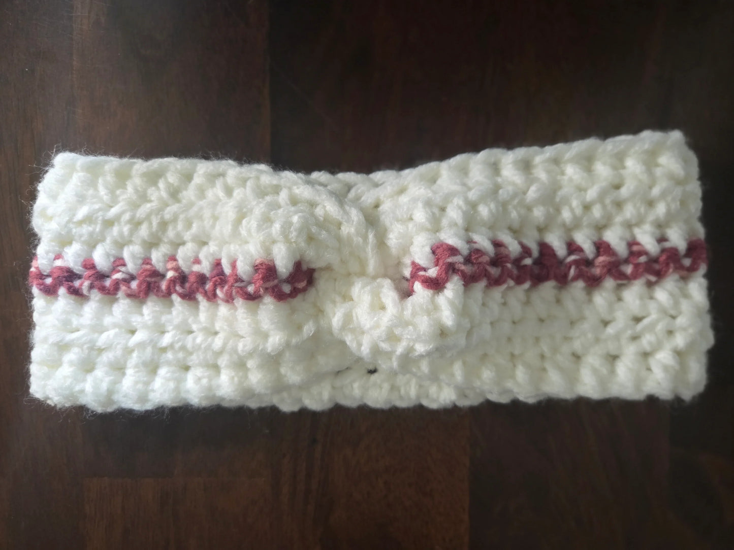 Handmade white crochet headband with pink stripe, knotted at center, on wooden table