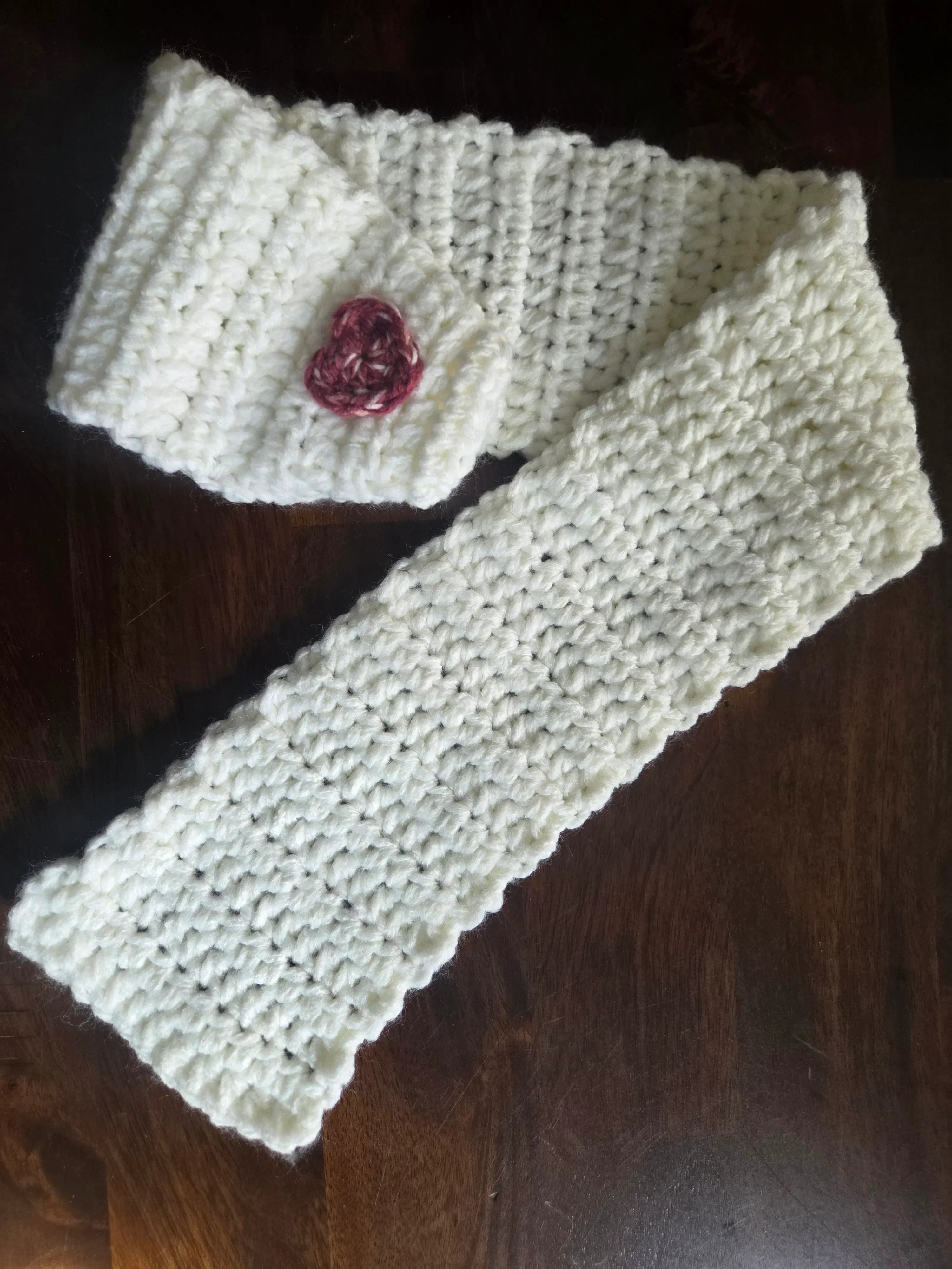 Handmade white crochet scarf with a small pink heart, displayed on a dark wood surface