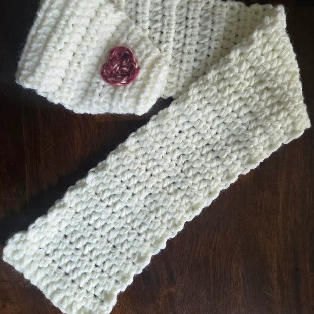 Handmade white crochet scarf with a small pink heart, displayed on a dark wood surface