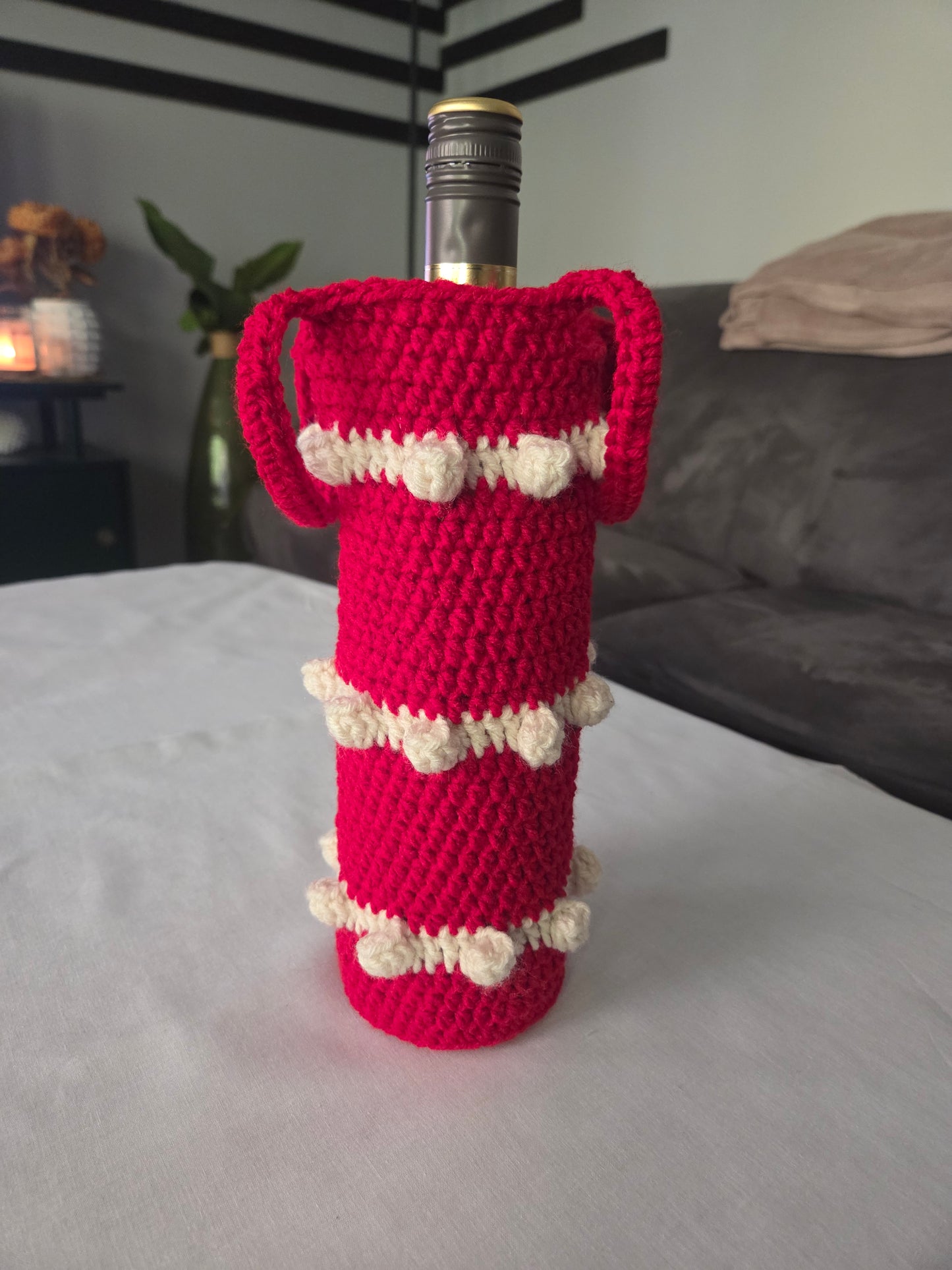 Red crochet wine bottle cover with white bobble accents on table