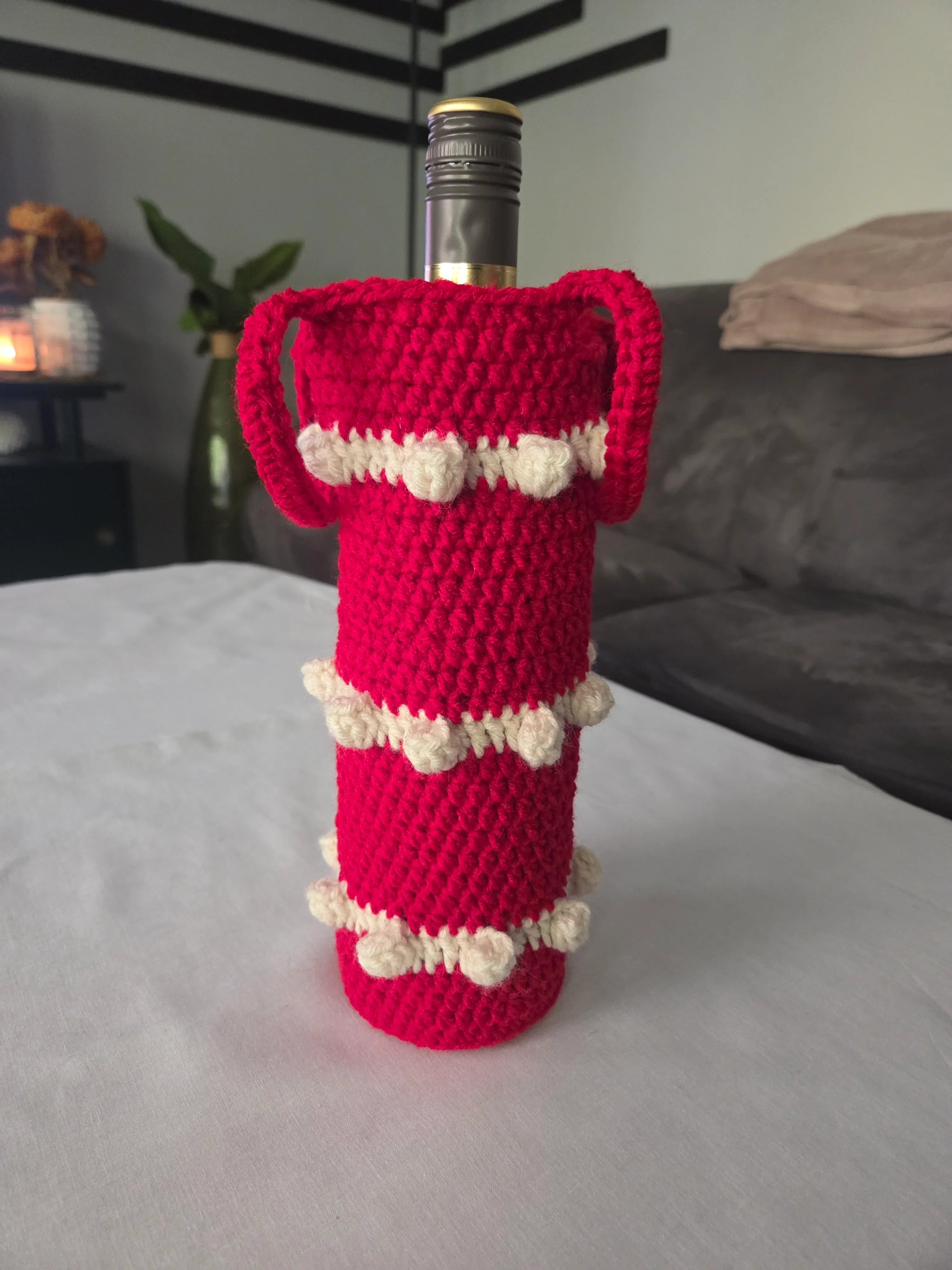 Red crochet wine bottle cover with white bobble accents on table