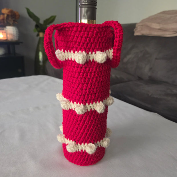 Red crochet wine bottle cover with white bobble accents on table