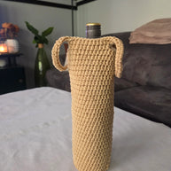 Beige crochet wine bottle cover with handles on a white table in a modern living room.
