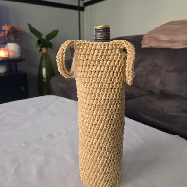 Beige crochet wine bottle cover with handles on a white table in a modern living room.