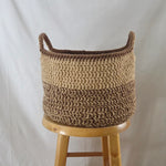 Handmade crochet basket with two-tone beige and brown yarn, featuring sturdy handles, displayed on a wooden stool.