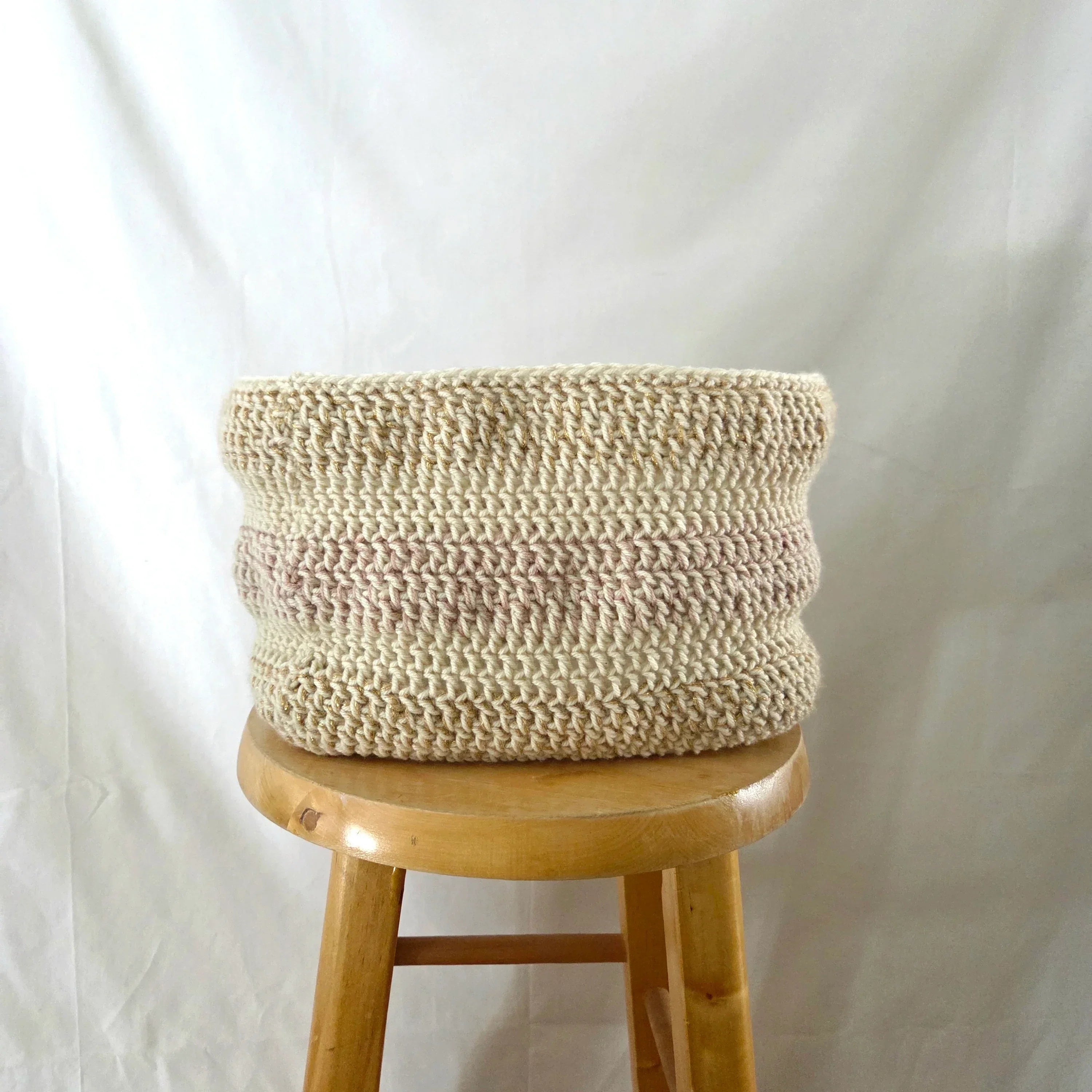 Handmade crochet basket in neutral tones displayed on a wooden stool against a white background