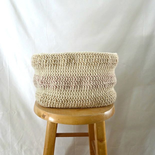 Handmade crochet basket in neutral tones displayed on a wooden stool against a white background