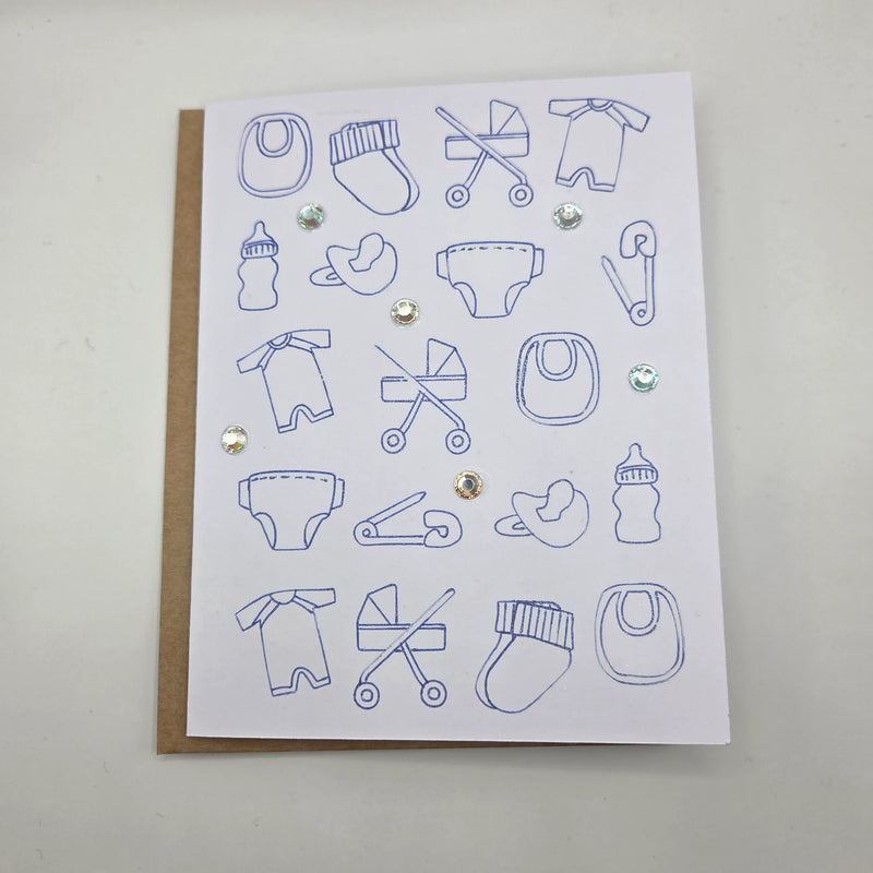 Handmade baby shower card with blue outline icons of onesies, strollers, bottles, and pacifiers, accented with glitter and rhinestones.
