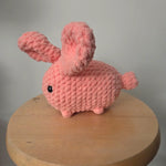 Handmade pink crochet bunny with black eyes, cozy amigurumi toy on wooden stand