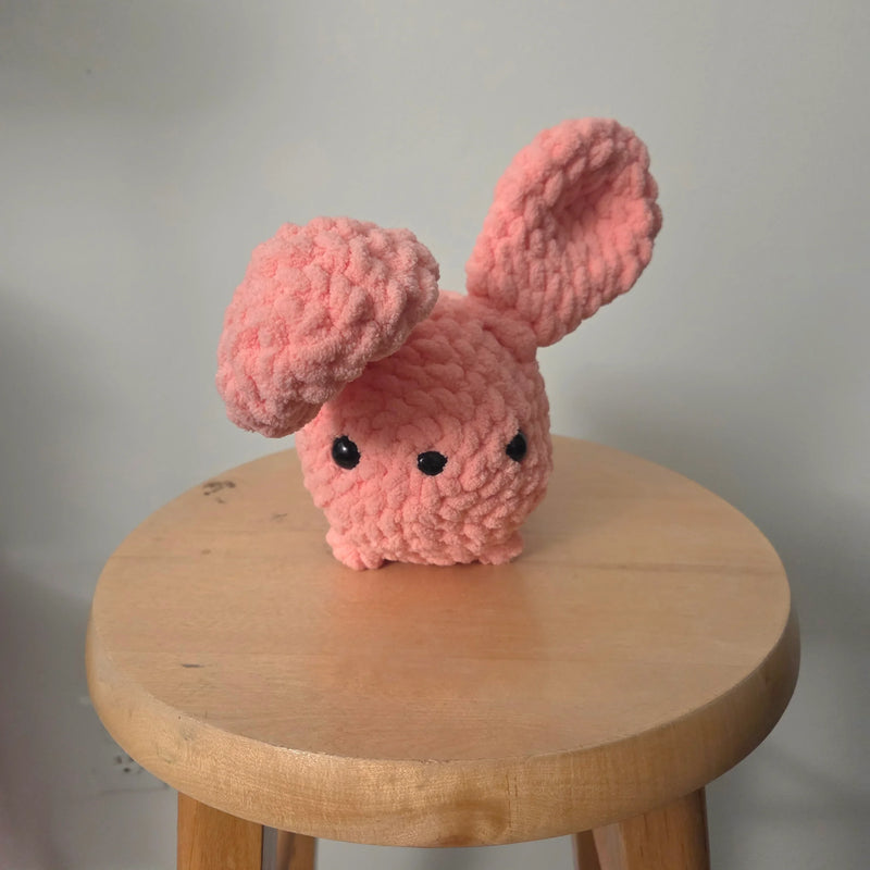Pink crochet bunny with floppy ears on wooden stool, handmade cozy decor