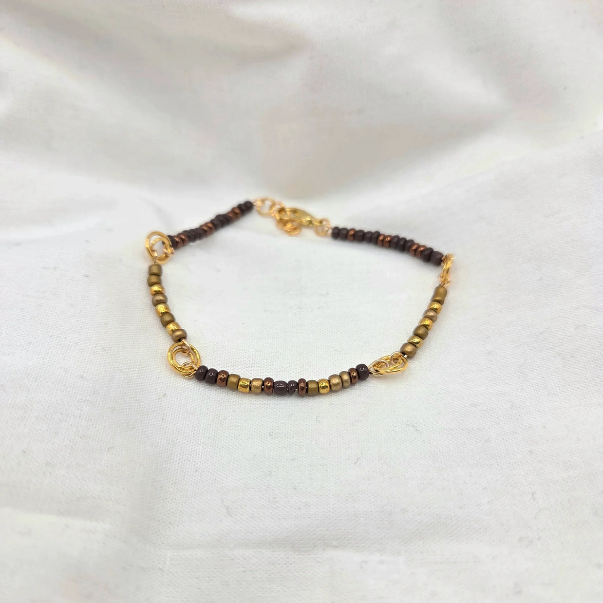Handmade beaded bracelet with gold, bronze, and brown beads on white fabric background