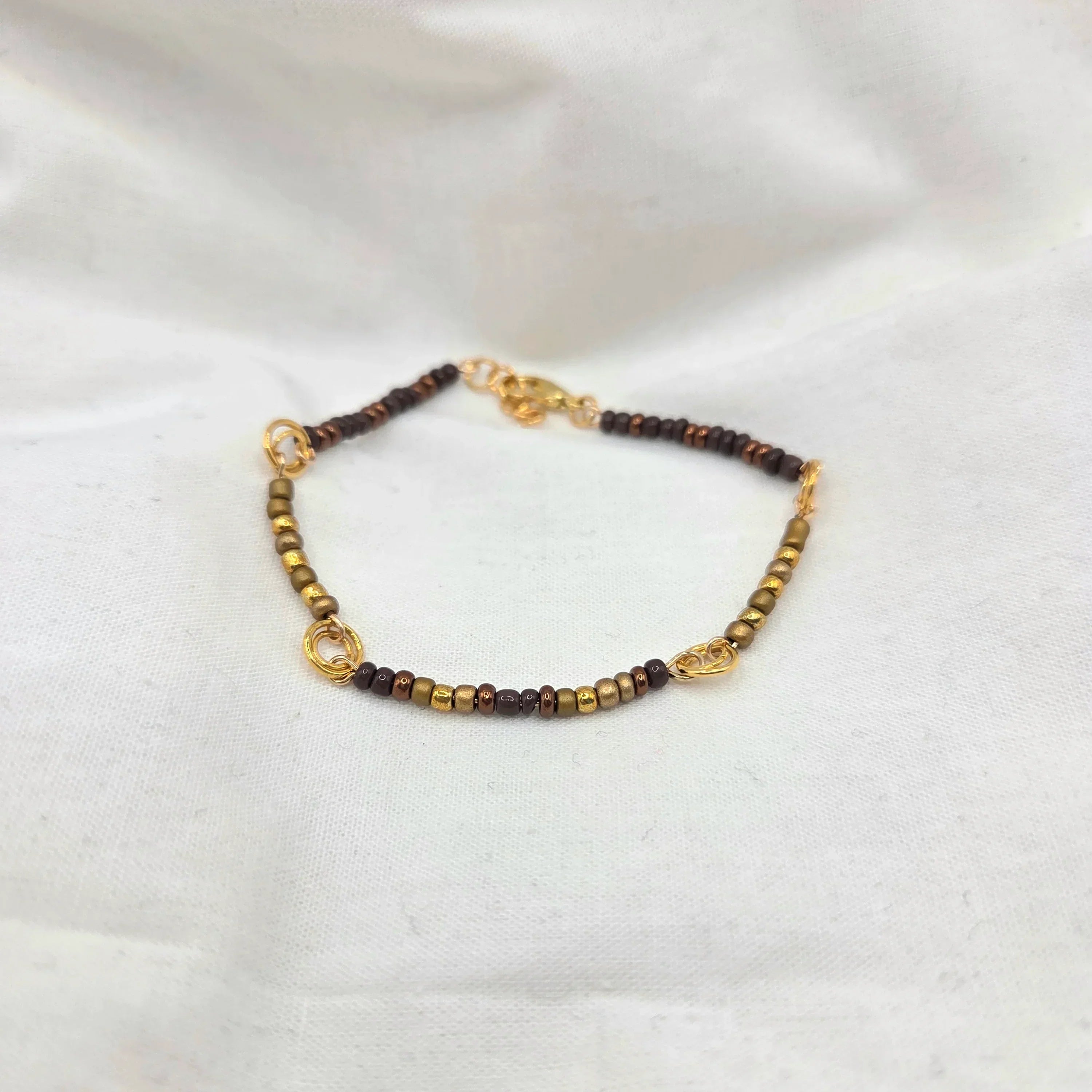 Handmade beaded bracelet with gold, bronze, and brown beads on white fabric background