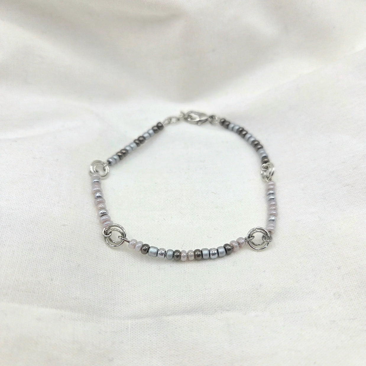 Delicate beaded bracelet with silver, gray, and pink beads, featuring interlocking rings on a soft white fabric background.