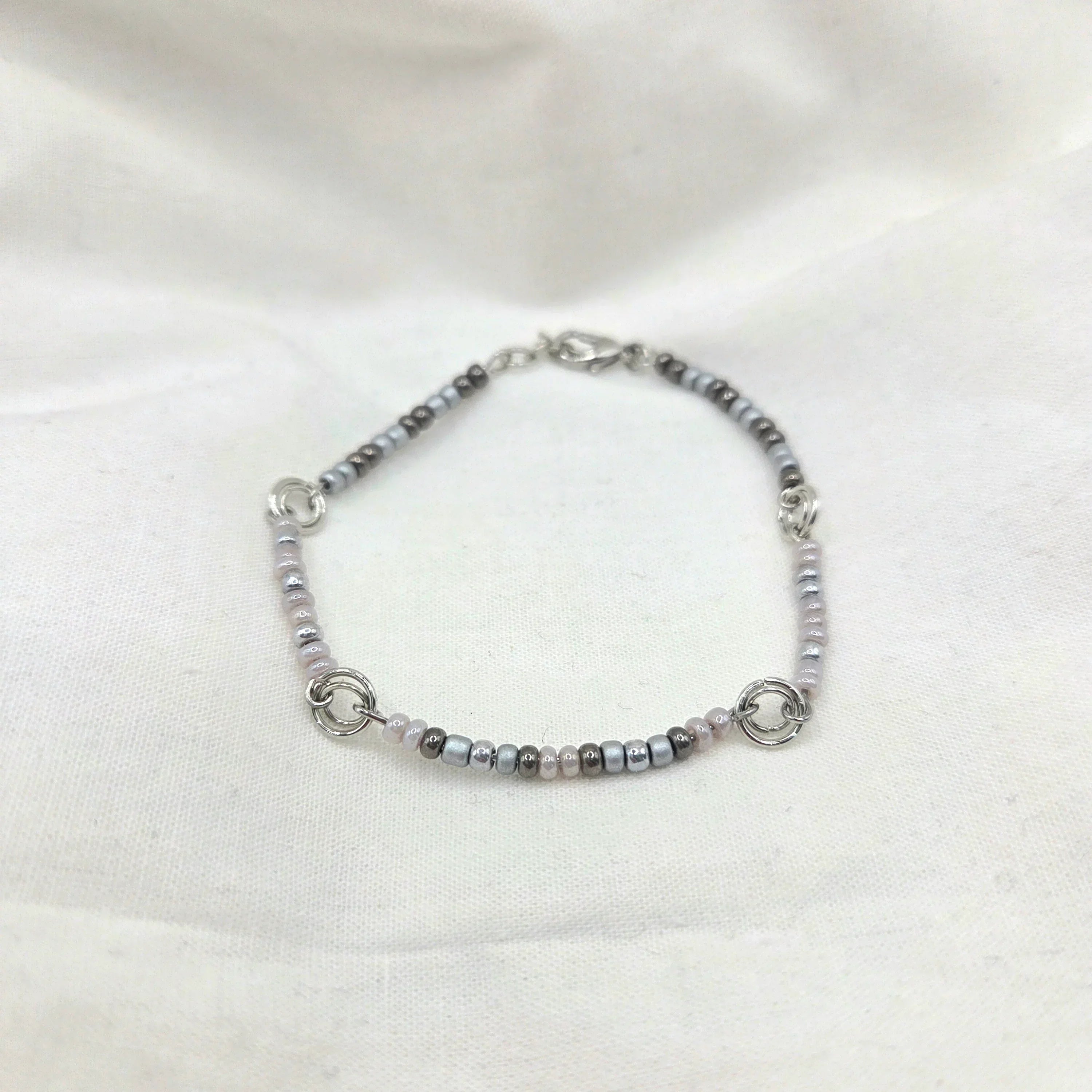 Delicate beaded bracelet with silver, gray, and pink beads, featuring interlocking rings on a soft white fabric background.