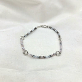Delicate beaded bracelet with silver, gray, and pink beads, featuring interlocking rings on a soft white fabric background.