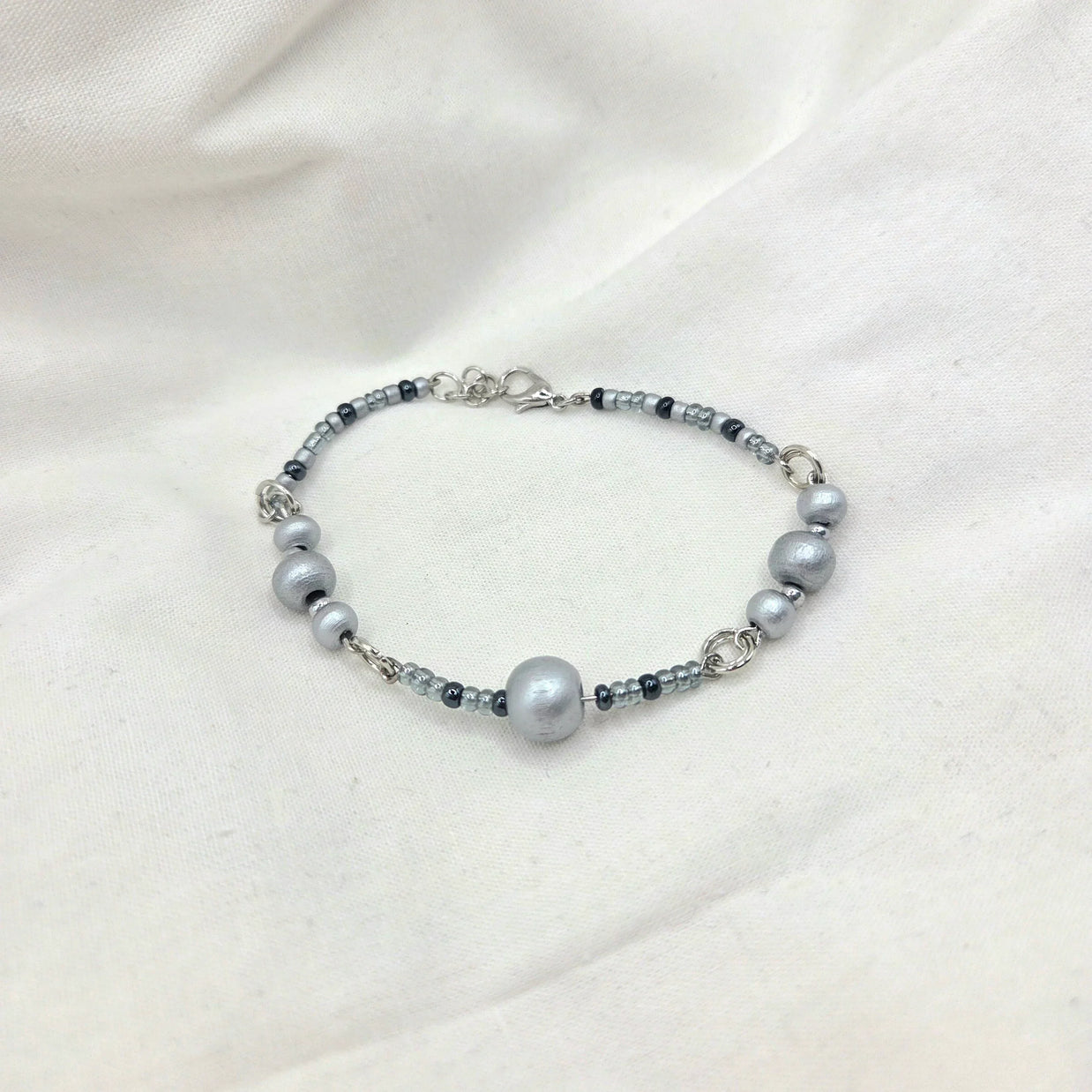 Gray beaded bracelet with silver-toned clasp, featuring matte and glossy beads on soft fabric background