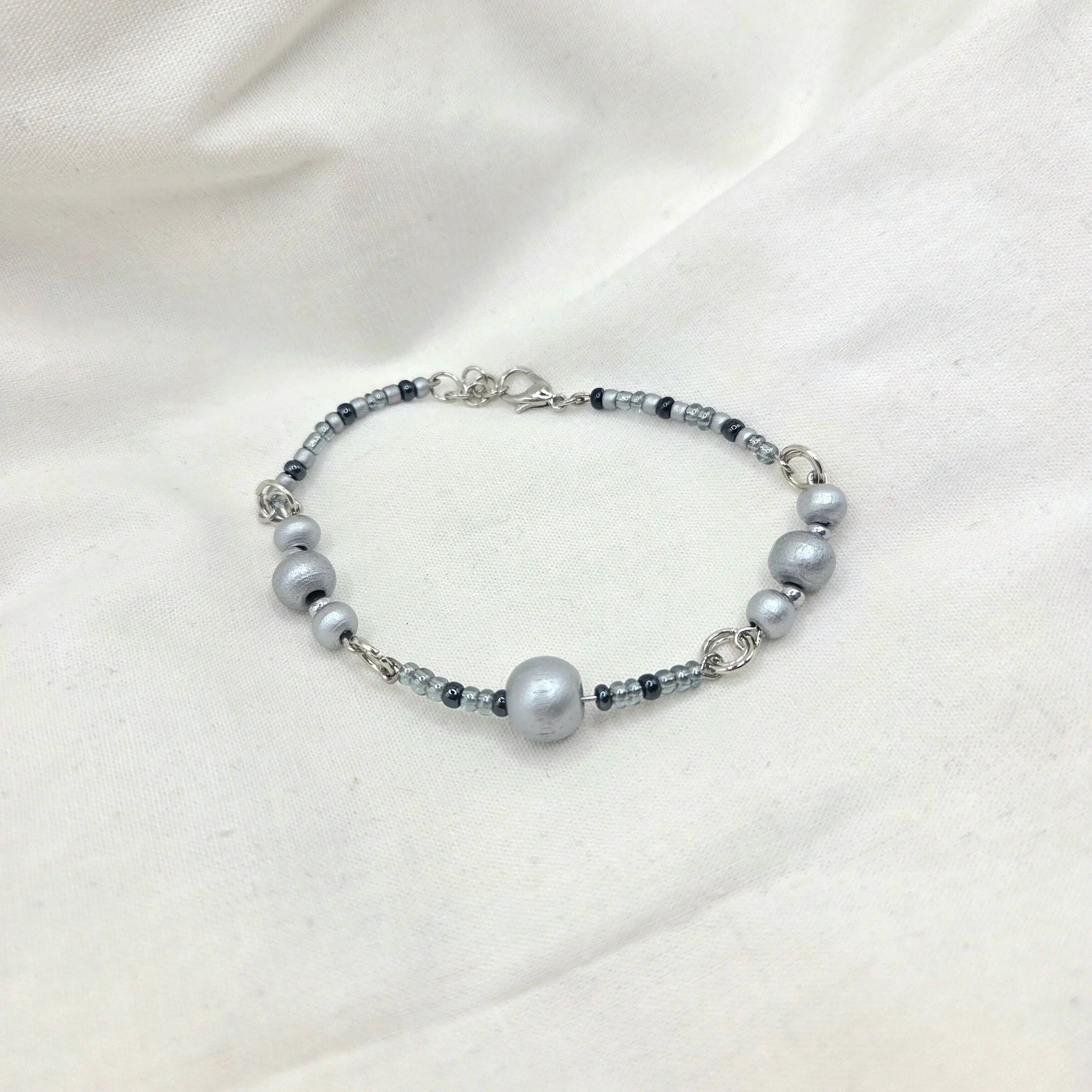 Gray beaded bracelet with silver-toned clasp, featuring matte and glossy beads on soft fabric background