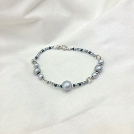 Gray beaded bracelet with silver-toned clasp, featuring matte and glossy beads on soft fabric background